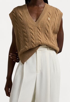 Gilet sans manches en maille torsadée marron à col en V, porté avec un pantalon blanc taille haute et large sur une personne à la peau foncée.