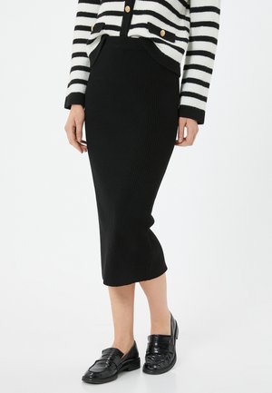Zwarte geribbelde kokerrok, knie-lengte, met een aansluitend ontwerp. Combinatie met zwarte gepatineerde loafers en een gestreepte cardigan.