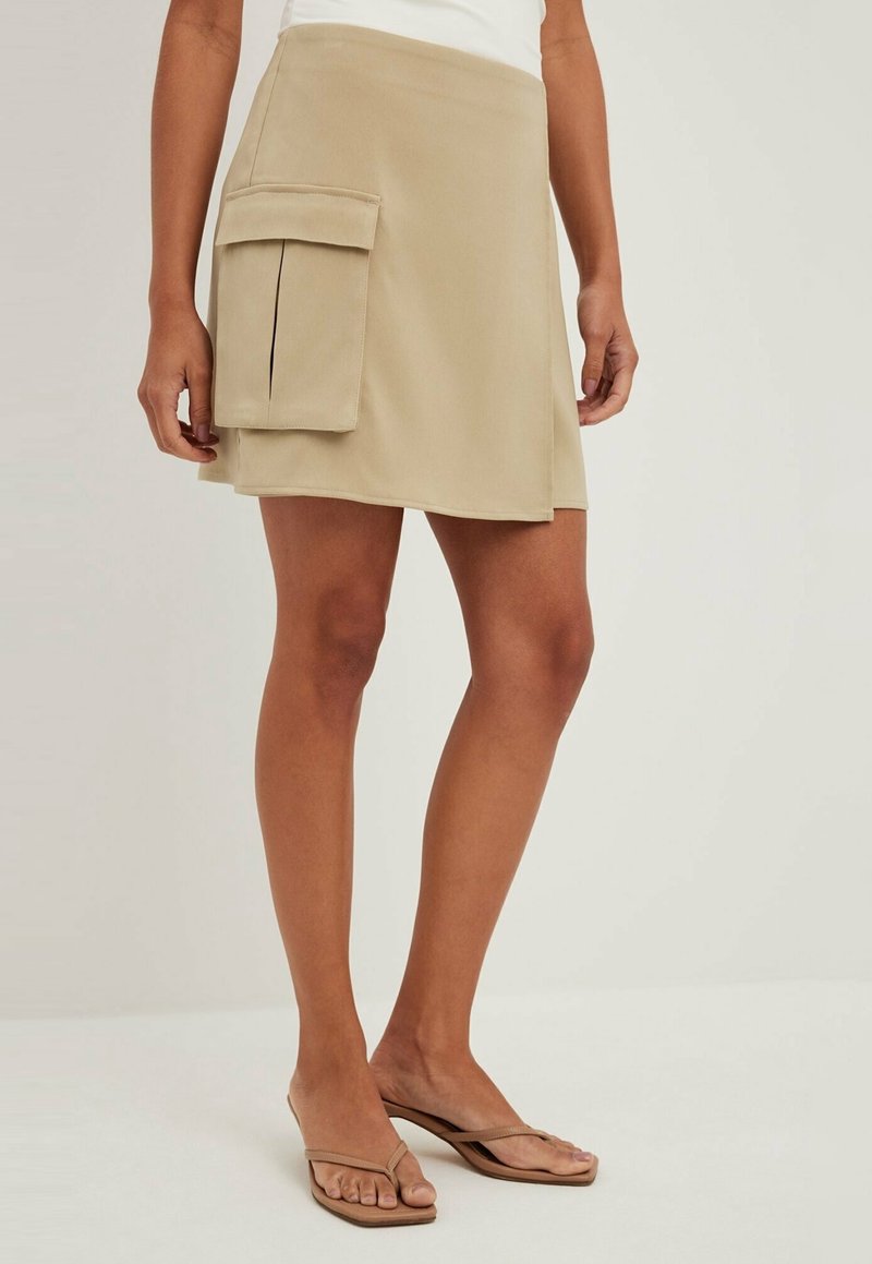 Femme portant une mini-jupe beige avec une grande poche latérale, un top blanc et des sandales beige type tongs, debout sur un fond uni.