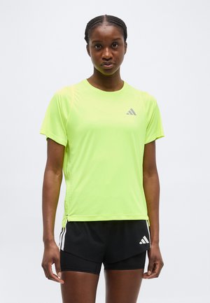 Γυναίκα που στέκεται φορώντας φωσφοριζέ κίτρινη μπλούζα Adidas και μαύρο σορτς Adidas με απλό φωτεινό φόντο.