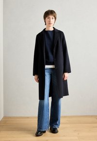 Manteau en laine noire avec de larges revers, deux boutons et des manches longues, superposé sur un pull bleu marine et un jean évasé bleu. Texture lisse.