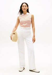 Skrzynkowy bezrękawnik w czerwono-białe paski, połączony z białymi spodniami o wysokim stanie. Modelka trzyma plecioną torbę ze słomy. Jasne tło.