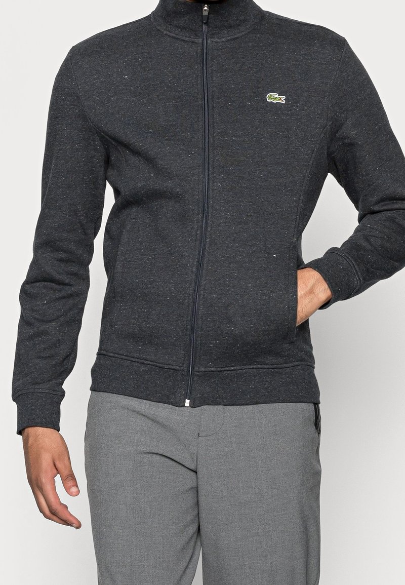Homme portant une veste zippée gris foncé avec un logo crocodile vert et un pantalon gris clair, une main dans la poche et l'autre le long du corps.