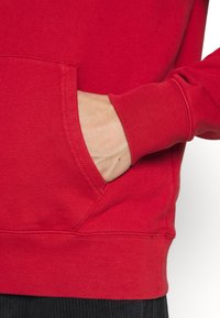 Main partiellement insérée dans la poche avant d'un sweat-shirt rouge à manches longues porté avec un pantalon noir.