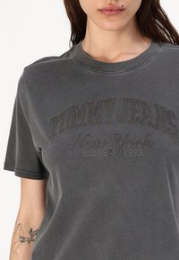 Graues Baumwoll-T-Shirt mit kurzen Ärmeln, das ein strukturiertes "TOMMY JEANS"-Logo, "New York" und "seit 1985" in kontrastierenden Fäden zeigt.