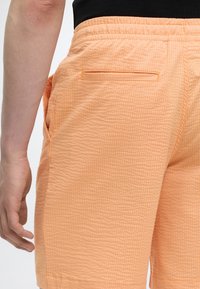 Κοντινό πλάνο ενός ατόμου που φορά πορτοκαλί υφασμάτινες βερμούδες με πίσω τσέπη και ελαστική μέση, με χέρι χαλαρό δίπλα στο σώμα.