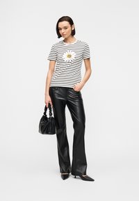 Γυναίκα που φοράει μαύρο δερμάτινο παντελόνι, ριγέ μπλουζάκι με σχέδιο μαργαρίτας, μαύρες μυτερές γόβες και κρατάει μια μαύρη τσάντα χειρός.