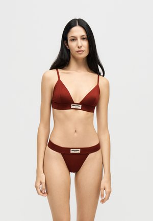 Femme aux longs cheveux foncés portant un ensemble bikini rouge foncé avec des bretelles fines et des étiquettes de marque sur le haut et le bas, sur un fond clair uni.