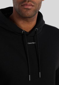 Primo piano di una persona che indossa un hoodie nero di Calvin Klein con i cordini visibili e il nome del marchio centrato sotto il colletto.