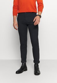 Tmavě modré slim-fit kalhoty vyrobené z strukturované tkaniny, s ohrnutými nohavicemi a předním zapínáním na knoflík, v kombinaci s černými kotníkovými botami.