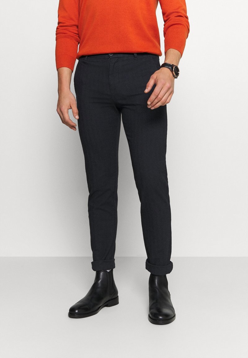 Tmavě modré slim-fit kalhoty vyrobené z strukturované tkaniny, s ohrnutými nohavicemi a předním zapínáním na knoflík, v kombinaci s černými kotníkovými botami.