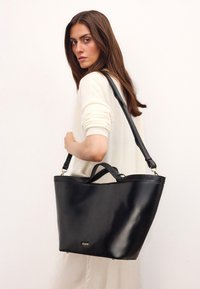 Black leather tote bag with a glossy finish, featuring dual handles and an adjustable strap. Includes gold brand logo detail.