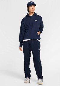 Felpa blu navy con tasca frontale e logo Nike bianco, abbinata a pantaloni della tuta blu navy. Indossata con un cappellino blu navy e sneakers beige.