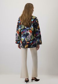 Giacca nera trapuntata con motivi colorati, che presenta teschi, foglie e forme geometriche. Abbinata a pantaloni a palazzo color crema e scarpe con tacco.