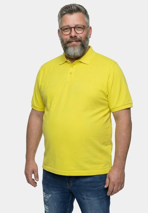 Mann mittleren Alters mit grauem Bart und Brille, der ein gelbes Poloshirt und blaue Jeans trägt, vor einem schlichten hellen Hintergrund stehend.