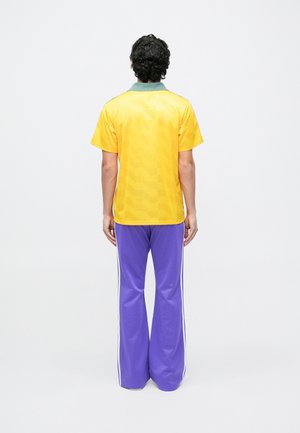 Mees lokkis juustega seisab seljaga vaataja poole, kandes kollast lühikese varrukaga särki ja lillasid alt laienevaid pükse valgete küljeribadega.