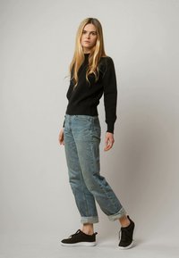 Schwarzer gerippter Pullover, hellblaue lockere Jeans mit umgeschlagenen Bündchen und dunkle Turnschuhe. Lange, glatte blonde Haare, stehende Pose vor einem grauen Hintergrund.