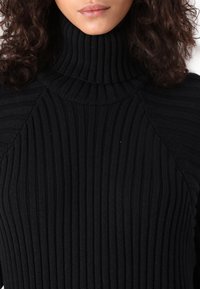 Pull noir à col roulé côtelé avec tissu texturé, doté d'un col haut et d'une silhouette ajustée, mettant en valeur des lignes verticales tricotées.