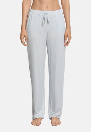 Kvinde iført lysegrå løstsiddende pyjamabukser med elastik i taljen og snøre, stående barfodet mod en ensfarvet baggrund.