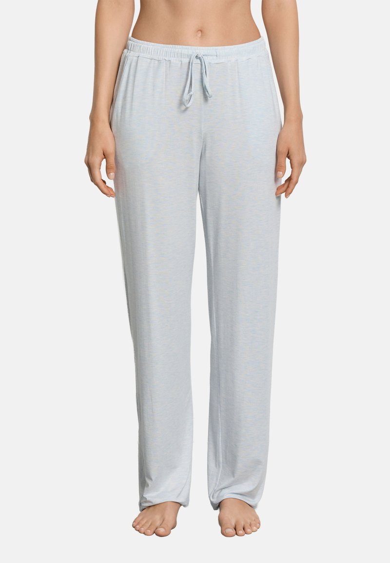 Kobieta nosząca luźne, jasnoszare spodnie do piżamy z elastycznym pasem i sznurkiem, stojąca boso na jednolitym tle.