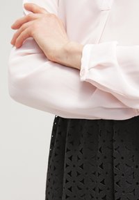 Helles rosa Blusenkleid mit langen, lockeren Ärmeln. Schwarzer Rock mit einem ausgeschnittenen Sternmuster. Knopfdetail am Ärmelbündchen sichtbar.