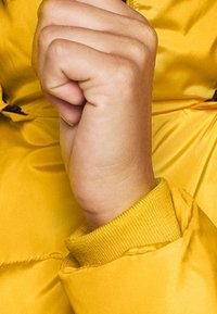 Gele donzige jackmanchet met geribbelde textuur en aangesloten pols. Hand in vuistpositie, met een gladde huid en natuurlijke kleur.