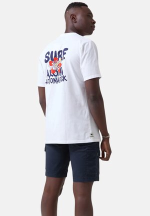Camiseta blanca de algodón con una flor colorida y el texto "SURF ALOHA" en la parte de atrás. Mangas cortas y escote redondo. Combinada con pantalones cortos azul marino.