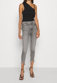 Black one-shoulder top, high-waisted grey jeans with frayed hems, and black strappy sandals. Model holds a small silver crossbody bag.