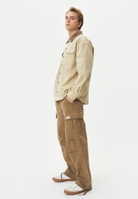 Beige langarmshirt met drukknoopjes en borstzak; bruine cargobroek met meerdere zakken; witte sneaker met bruine zolen.