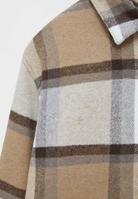 Tissu en laine à carreaux beige, marron et blanc avec une texture douce, montrant un col et une partie d'une manche sur un vêtement.