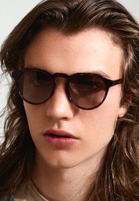 Gafas de sol redondas de carey con lentes oscuros, montura gruesa y un detalle distintivo en el puente. Textura suave, diseño moderno.