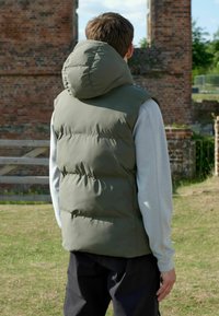 Olivgrüne gepolsterte Weste mit hoher Kapuze und gesteppten Abschnitten. Über einem hellgrauen Langarmoberteil getragen. Schlichtes, funktionales Design.