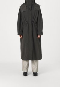 Parka long, vert foncé avec un col haut, un cordon de serrage à l'avant et des poches surdimensionnées. Porté sur un pantalon léger à motifs texturés et des bottines.