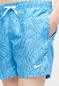 Zili peldēšanas šorti ar melniem abstraktiem vilkmes rakstiem, balta aukla un balts Nike logotips uz kreisās kājas, roka labajā kabatā.