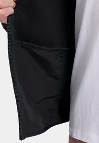 Mano sosteniendo una chaqueta negra abierta que muestra el suave tejido interior y el bolsillo, junto a una persona que lleva una camisa blanca.