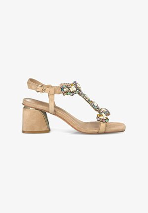 Sandale à talon en suède beige avec un talon bloc et des brides décoratives ornées de bijoux, présentant des pierres multicolores et un design à lanière en T.