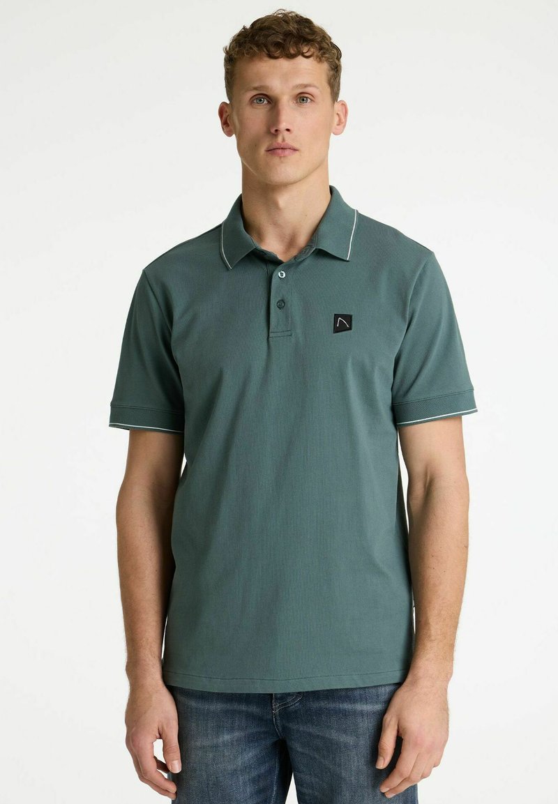 Jeune homme portant un polo vert à manches courtes avec un bord blanc, debout devant un fond blanc uni, les mains détendues le long du corps.