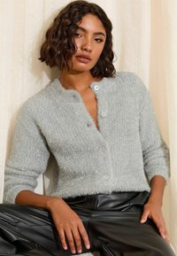 Gaiši pelēks, pūkains kardigāns ar apaļu kakla izgriezumu, teksturētu ribojumu un baltām pogām. Apģērbs kombinēts ar melnām ādas biksēm.