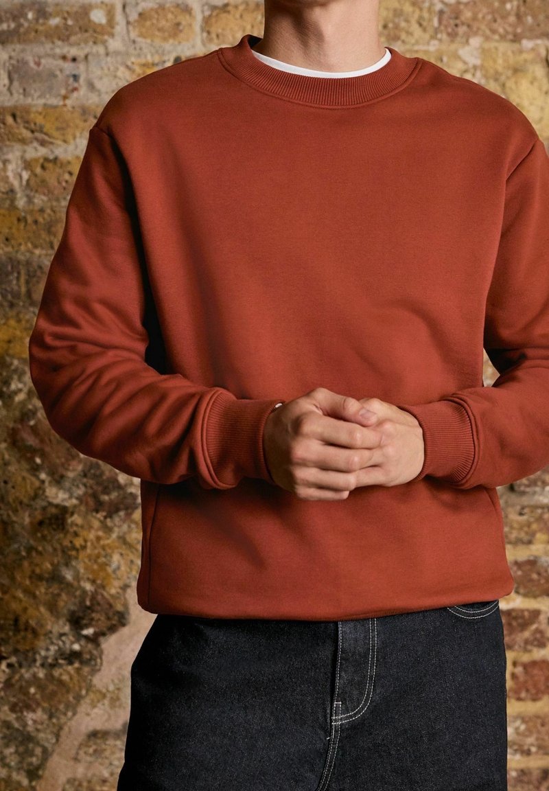 Personne portant un sweat-shirt couleur rouille et un jean foncé, les mains jointes devant, debout devant un mur de briques texturé.