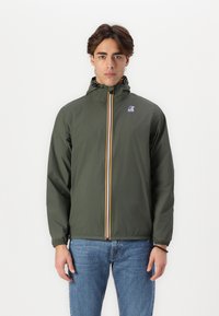Chaqueta verde oliva con capucha, cremallera vertical amarilla y azul, puños elásticos y un pequeño logotipo en el pecho. Lleva puesta con jeans azules.
