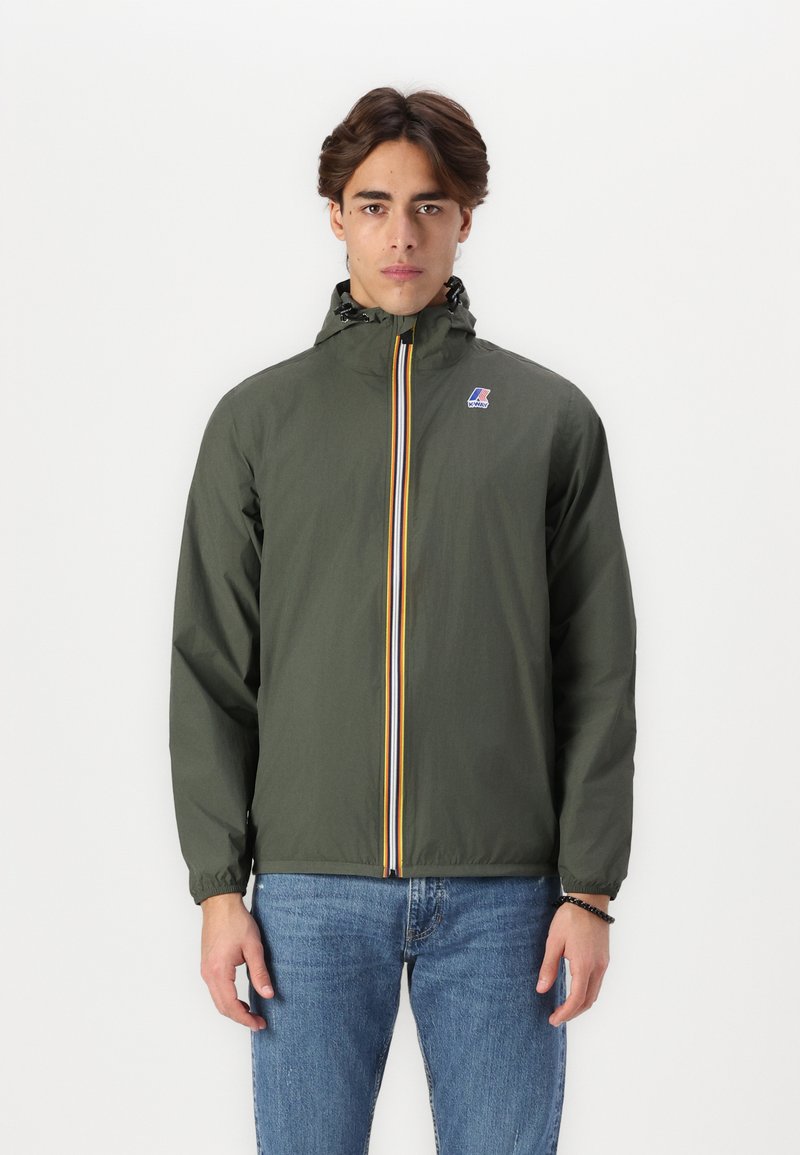 Chaqueta verde oliva con capucha, cremallera vertical amarilla y azul, puños elásticos y un pequeño logotipo en el pecho. Lleva puesta con jeans azules.