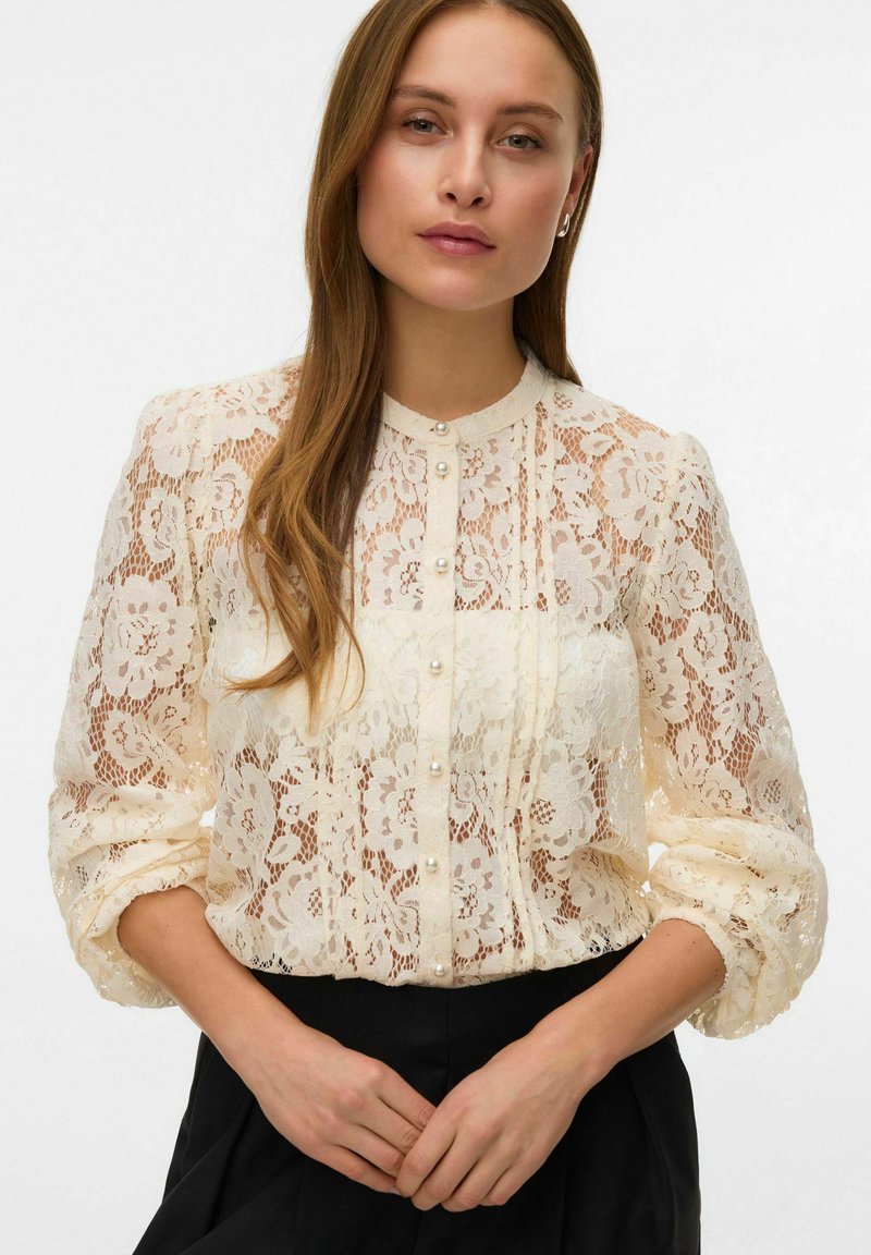 Koronkowa bluzka w kolorze kremowym z kwiatowym wzorem, z zapięciem na guziki i guzikami przypominającymi perły. Posiada bufiaste rękawy oraz okrągły kołnierzyk.