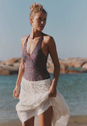 Joven mujer con el cabello recogido en un moño lleva un traje de baño estampado y una falda blanca de encaje, de pie junto a la costa rocosa bajo un cielo despejado.