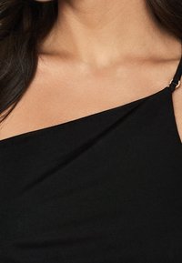 Top noir en tissu côtelé avec un décolleté asymétrique et une seule sangle ornée d'un détail en métal doré. Texture lisse et coupe ajustée.