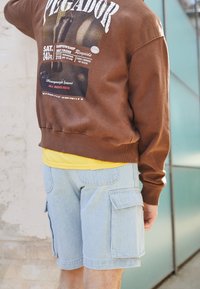 Hnědá mikina s grafickým potiskem; žluté tričko pod tím. Světle modré cargo šortky s bočními kapsami. Hladká textura a ležérní design.