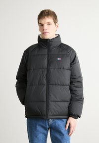 Zwarte donsjas met een hoge kraag, rits aan de voorkant, horizontale naden en een klein logo op de linkerborst. Draagt met blauwe jeans.