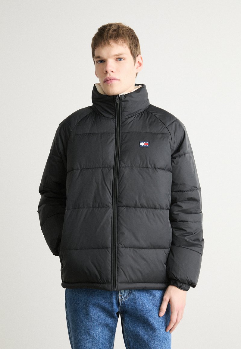 Zwarte donsjas met een hoge kraag, rits aan de voorkant, horizontale naden en een klein logo op de linkerborst. Draagt met blauwe jeans.