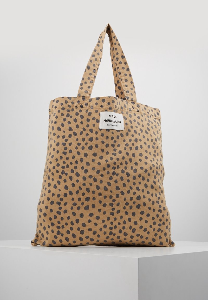 Beige tygväska med svarta oregelbundna fläckar och tyghandtag, med en vit etikett som läser "Mads Nørgaard Copenhagen."