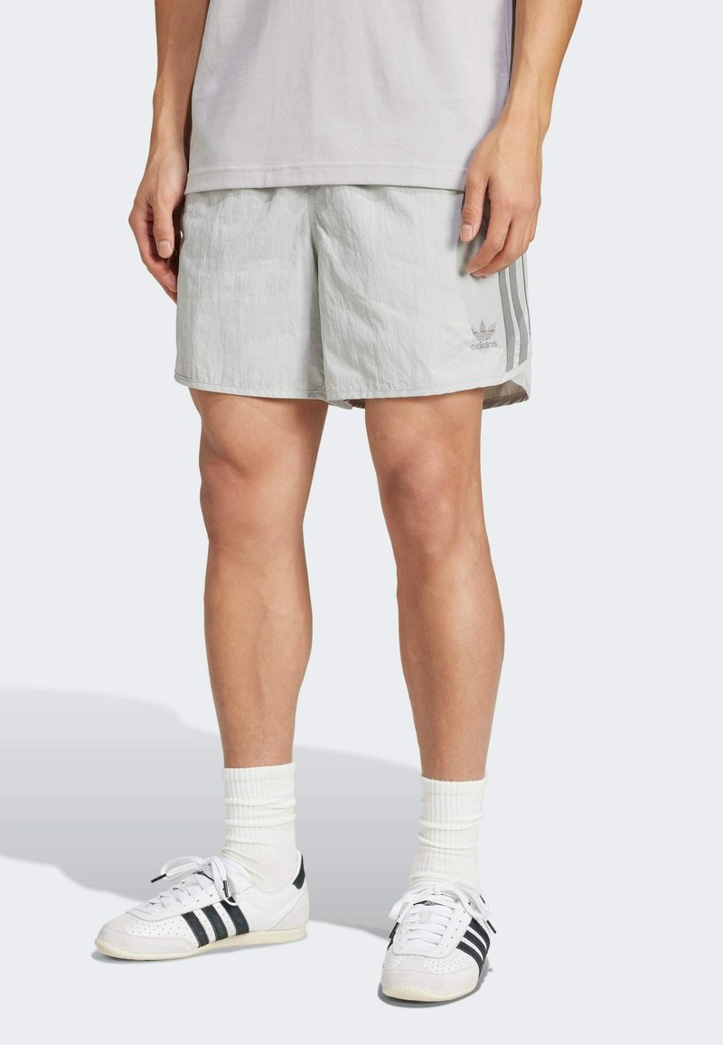 Pantalones cortos deportivos de color gris claro con una cinturilla elástica, que cuentan con tres rayas verticales a los lados y un logo en la parte frontal.