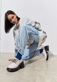 Încălțăminte bej deschis cu toc gros negru, având șireturi negre. Modelul poartă blugi de denim de culoare albastră deschisă și un pulover gri cu model.